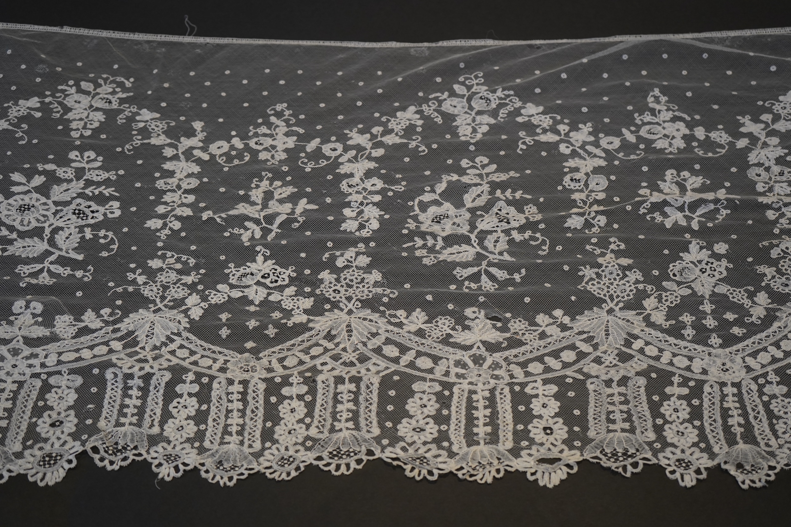 A deep elaborately designed flounce of mid 19th century Brussels bobbin appliquéd lace on net, with finished lace ends, worked with an intricate floral border below large floral cartouches framed in similar motifs and al